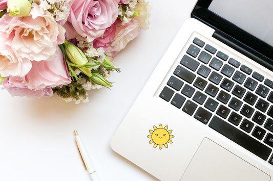A flat lay of a clean workspace showing a laptop with a cute smiling sun sticker on it, a bouquet of pink and white flowers, and a white pen.
