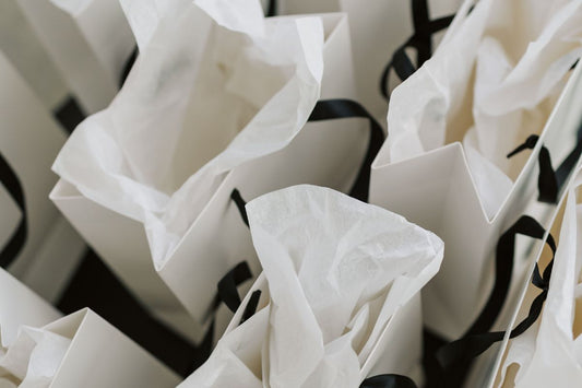 A close-up shot of several white paper gift bags with black ribbon handles, filled with delicate, crinkled white tissue paper
