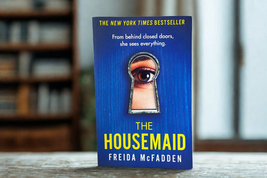A photo of the paperback cover of the thriller "The Housemaid" by Freida McFadden. The blue cover features a large keyhole with an eye looking through it, and the book is standing on a light-colored, buttoned cushion.