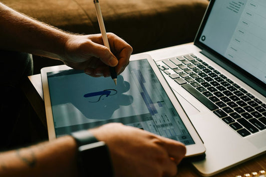 A person drawing a logo on a tablet with a stylus, placed next to a laptop