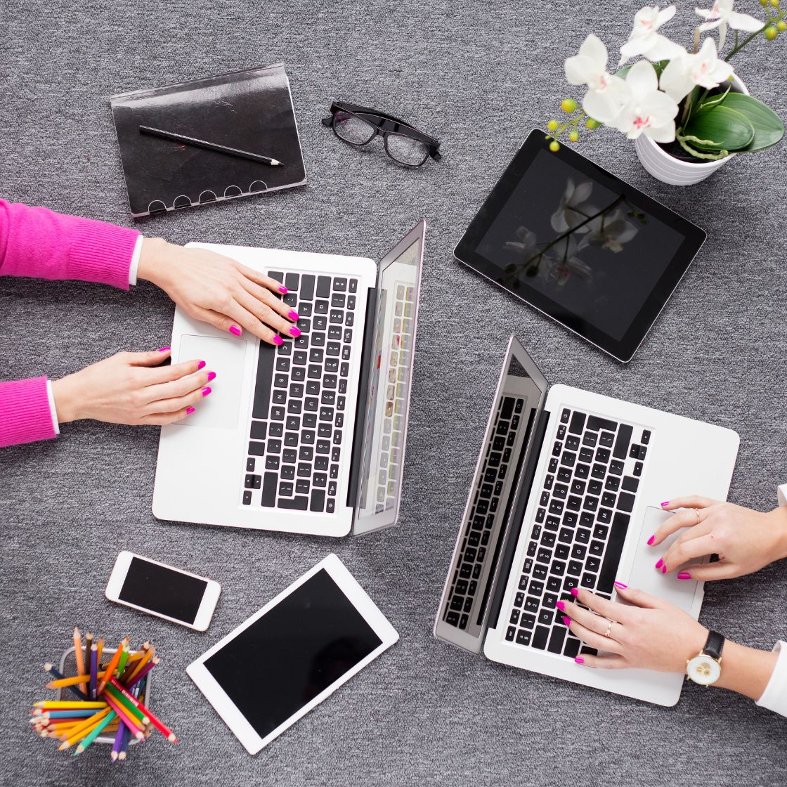 Two people using laptops on a gray desk with tablets, smartphone, and stationery.