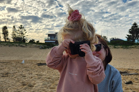 A candid photo of a daughter and working mum enjoying a moment on the beach, the little girl is pretending to take a photo with a camera.