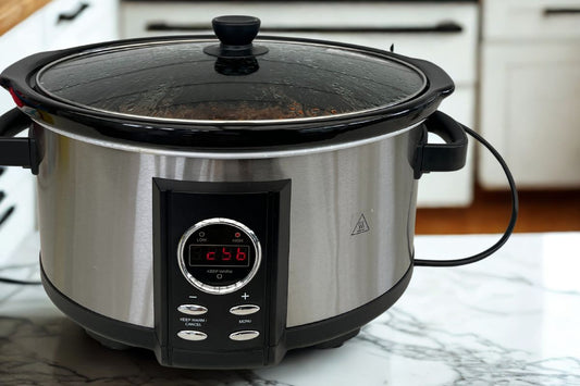 This image shows a Kmart 6.5L slow cooker, which appears to be a stainless steel appliance with black trim and a glass lid. The slow cooker has a digital display (showing 1:42) and several control buttons on its front panel.