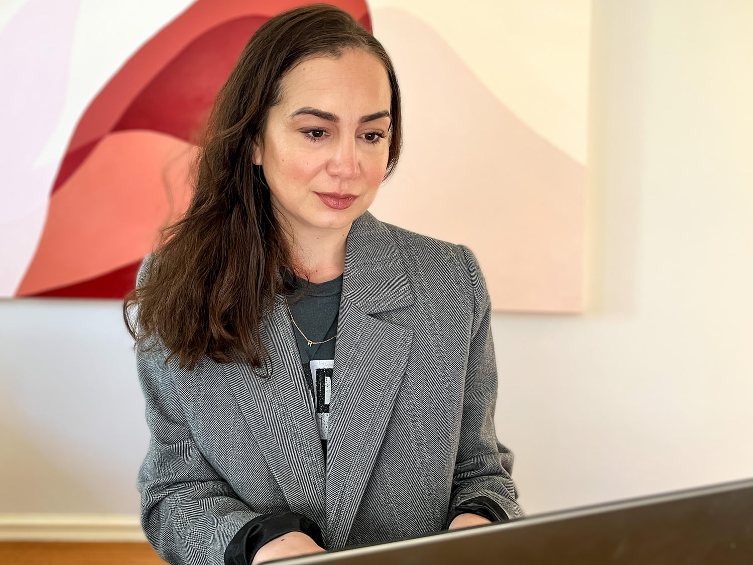 A focused SEO freelancer in Sydney with long brown hair, wearing a gray blazer over a dark shirt, works on a laptop at a wooden table. The background features a modern abstract painting in shades of red, pink, and beige on a white wall, creating a professional yet creative workspace